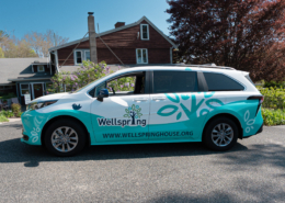 Side view of Wellspring House’s new hybrid passenger van with custom teal-and-white wrap and logo, parked in front of Wellspring House in Gloucester, MA.