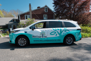 Side view of Wellspring House’s new hybrid passenger van with custom teal-and-white wrap and logo, parked in front of Wellspring House in Gloucester, MA.
