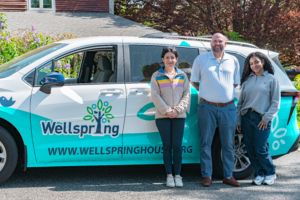 Three staff from Wellspring's Homelessness Prevention program in front of the newly wrapped Wellspring hybrid passenger van, smiling during the ribbon-cutting celebration.